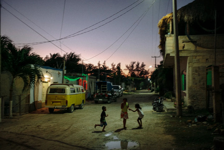 Children playing under the glow of streetlights in El Cuyo, Mexico, while the sky blends into warm night hues, 2019.